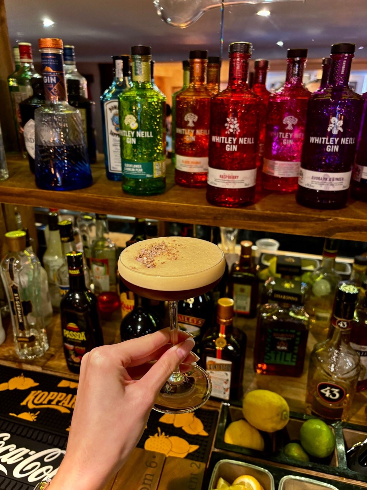 An espresso martini in front of the gin shelf at the bar